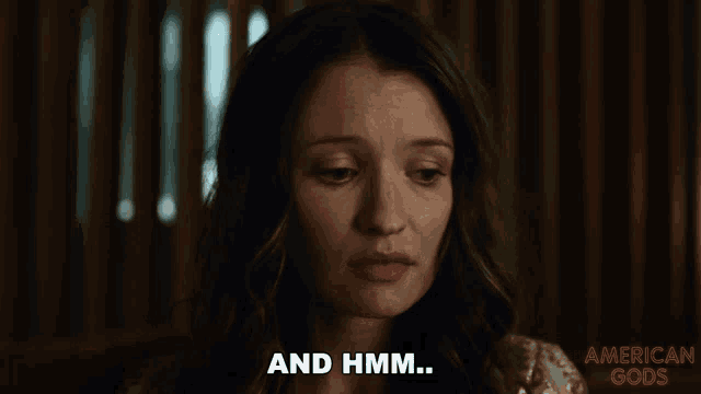 a woman says " and hmm " in front of an american gods poster