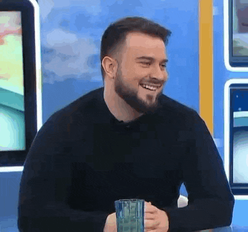 a man with a beard is smiling while sitting at a table with a glass of water .