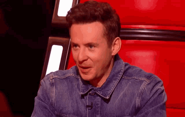 a man in a denim jacket is sitting in a red chair and making a funny face .