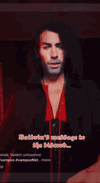 a man with long hair is wearing a black jacket and a red shirt and has a message on his face .