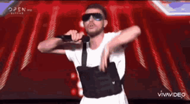 a man wearing sunglasses and a vest is singing into a microphone on a stage .