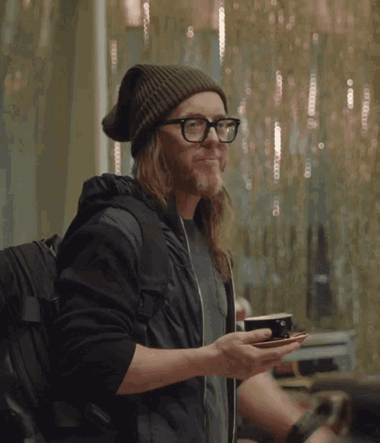 a man with long hair and glasses holds a cup of coffee