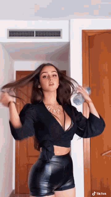 a woman in a black crop top and leather shorts is standing in a room holding her hair .