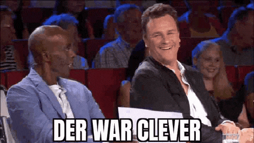 two men are sitting next to each other in a theatre and laughing while a crowd watches .