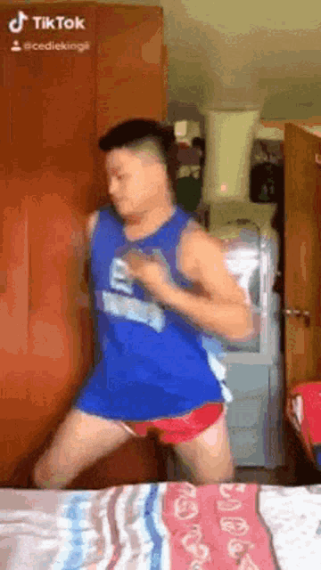 a man in a blue tank top and red shorts is dancing on a bed in a bedroom .