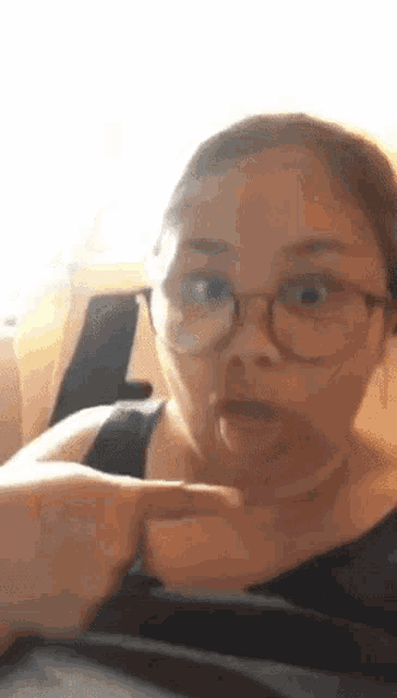 a woman wearing glasses is making a funny face with her tongue out and pointing at her chest .