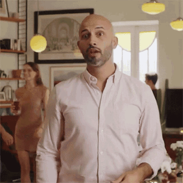a bald man in a pink shirt is standing in a living room with a woman in a dress behind him .