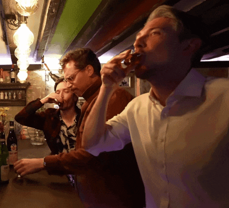 a man drinking from a shot glass with a bottle of wine in the background