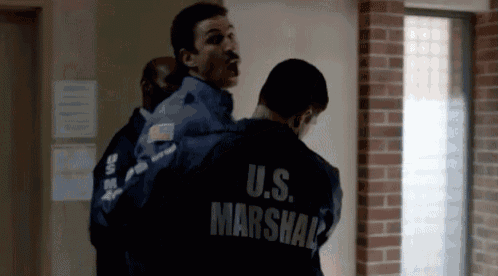 a man wearing a u.s. marshal jacket is being escorted by another man