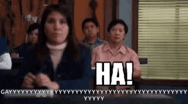 a group of people are sitting in a room and a woman is standing in front of them and says `` ha ! ''