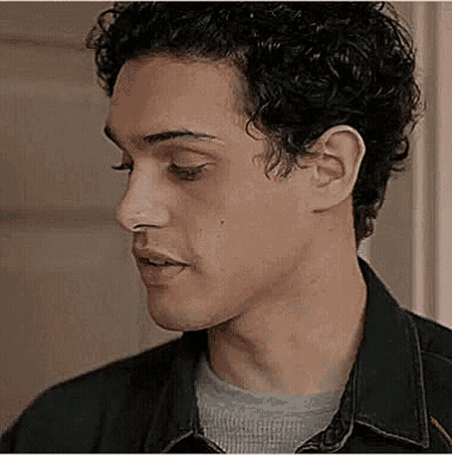 a close up of a man 's face with curly hair wearing a denim jacket and a grey shirt .