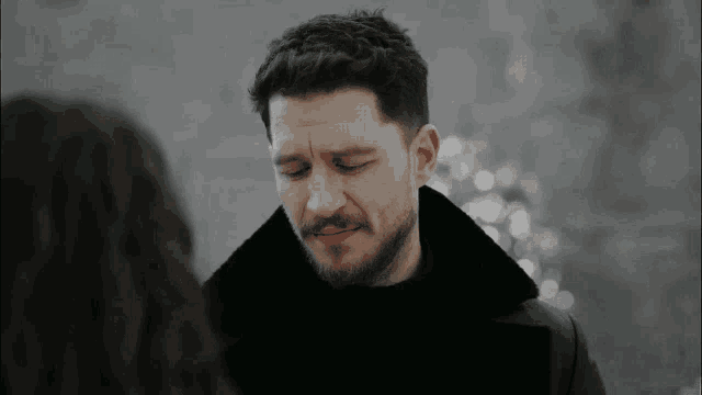 a man with a beard and a black coat is looking at a woman with his eyes closed .