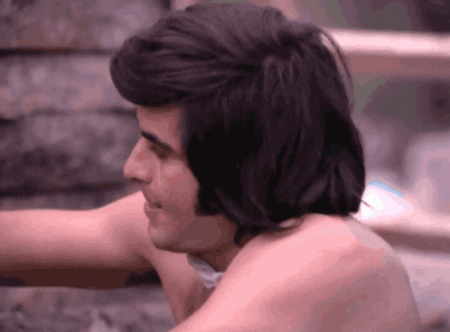 a shirtless man with long dark hair looks to his right