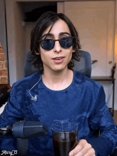 a young man wearing sunglasses and a blue shirt is sitting in a chair holding a glass of soda .