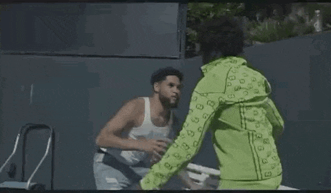 a man is drinking water from a plastic bottle while another man stands next to him on a park bench .