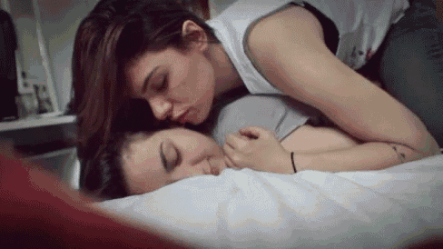two women are laying on a bed , one is kissing the other .