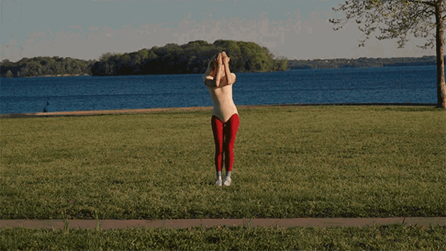 a woman in red pants and a white top stands in a grassy field near a body of water