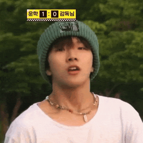 a young man wearing a green beanie and a white shirt is standing in front of trees .