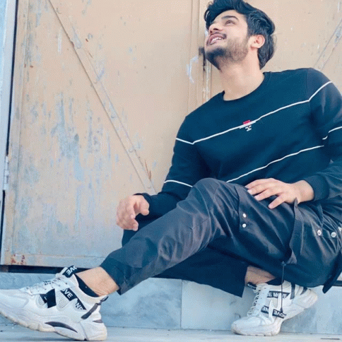 a man wearing a black sweater and black pants sits on a ledge with his legs crossed