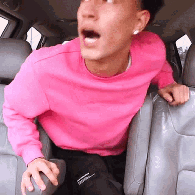a man in a pink sweater is sitting in the back seat of a car with his mouth open .