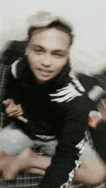 a man in a black hoodie is sitting on a bed with his legs crossed .