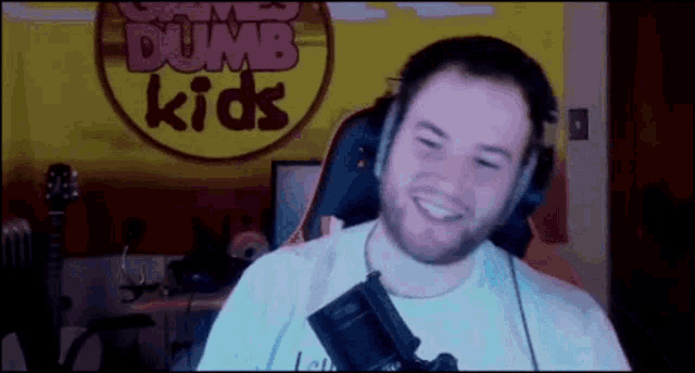 a man wearing headphones and a white shirt is smiling in front of a sign that says dumb kids .