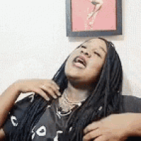 a woman with braids is sitting on a couch with her mouth open and a picture of a woman on the wall behind her .