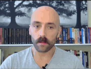 a bald man with a beard is standing in front of a bookshelf with a picture of trees on it