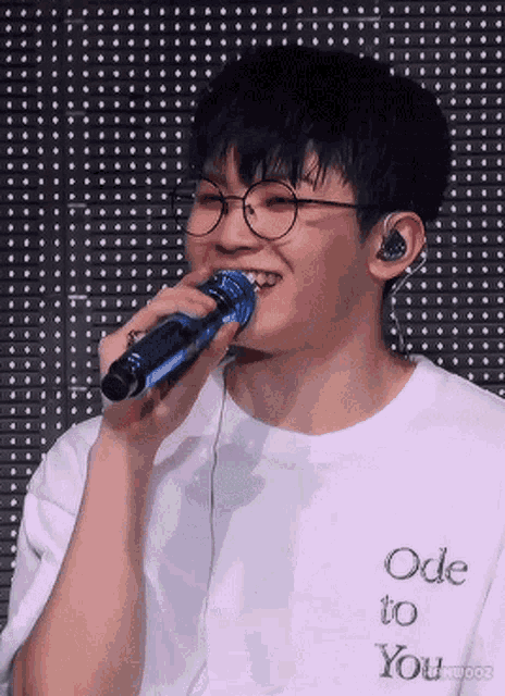 a young man wearing glasses is singing into a microphone while wearing a white shirt that says ode to you .