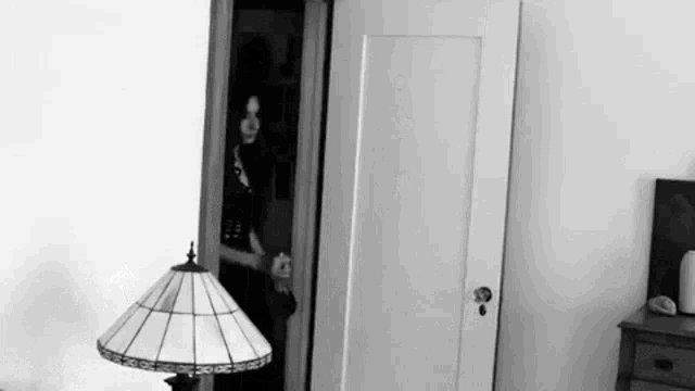 a black and white photo of a person standing in a doorway next to a lamp in a bedroom .