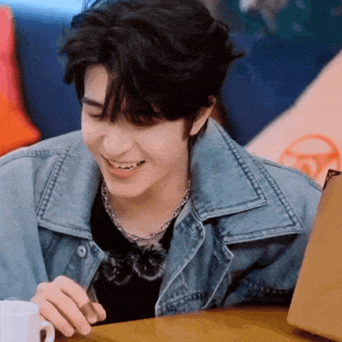 a young man in a denim jacket is sitting at a table with a cup of coffee and a laptop .
