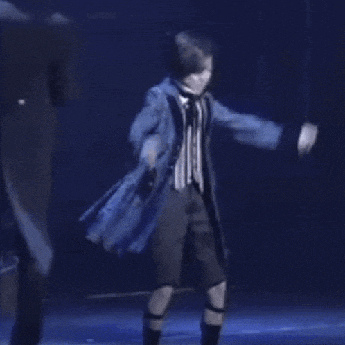 a young boy is dancing on a stage in a blue coat and shorts .