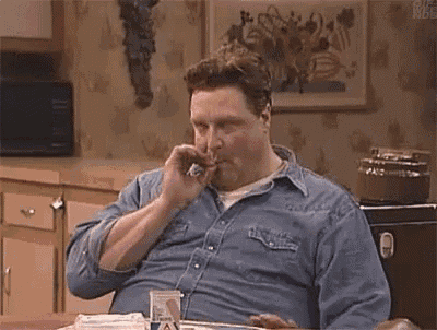 a man in a denim shirt is sitting at a table eating a hamburger and drinking beer .