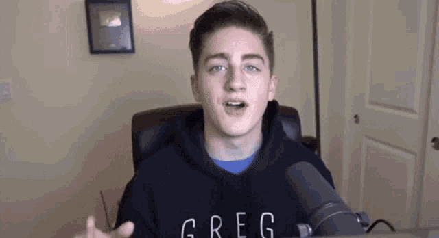 a young man is sitting in a chair in front of a microphone wearing a hoodie that says greg .