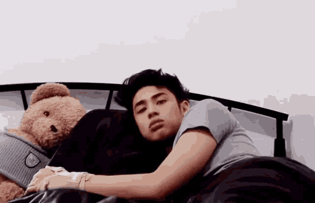 a young man is laying on a bed with a teddy bear next to him .