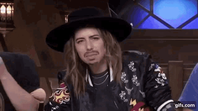 a man with long hair and a beard is wearing a hat and jacket .