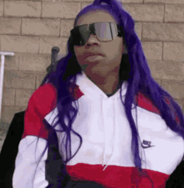 a woman with purple hair and sunglasses is wearing a red , white , and black nike sweatshirt .