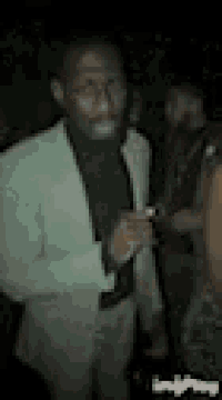 a man in a suit is standing in a dark room holding a cigarette .