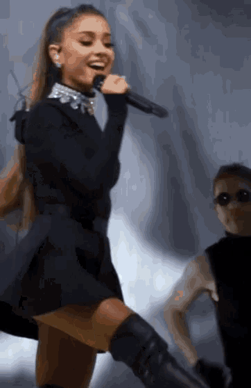a woman in a black dress is singing into a microphone with a man in sunglasses behind her