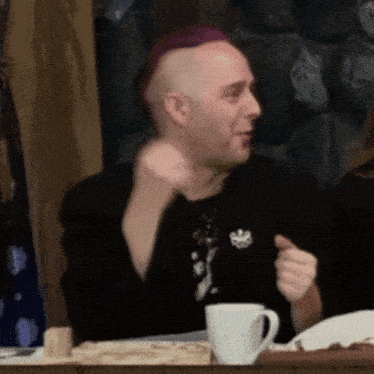 a man wearing a purple mohawk is sitting at a table with a cup of coffee