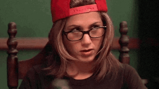 a woman wearing glasses and a red hat is sitting in a chair holding a cucumber .