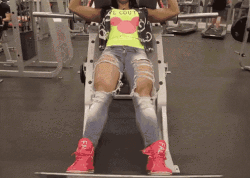 a woman wearing a mickey mouse shirt is squatting on a machine in a gym