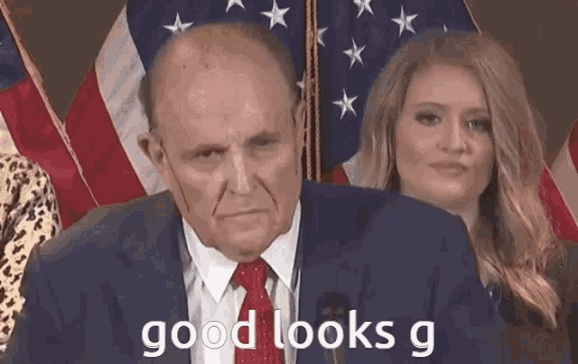 a man in a suit and tie is standing in front of an american flag while a woman looks on .