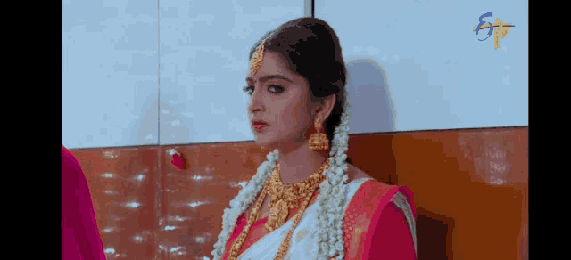 a woman in a pink and white saree and gold jewelry is standing in front of a wall .