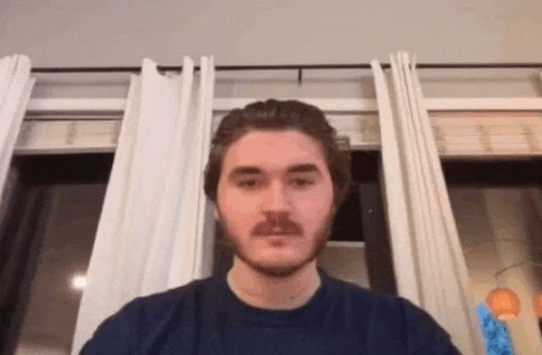 a man with a beard and mustache is sitting in front of a window with curtains .