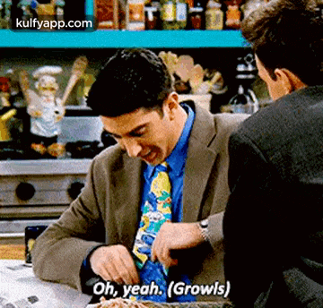 a man in a suit and tie is talking to another man in a kitchen while cutting spaghetti .