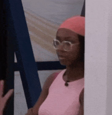 a woman wearing glasses and a headband is standing next to a wall and looking at the camera .