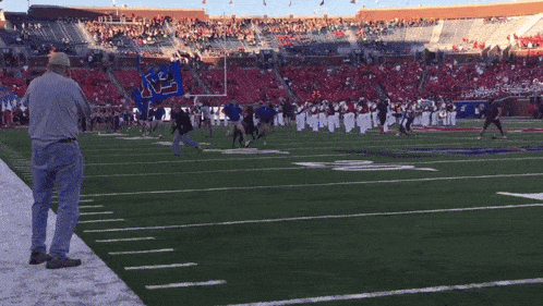 a man stands on the sideline of a football field watching a marching band