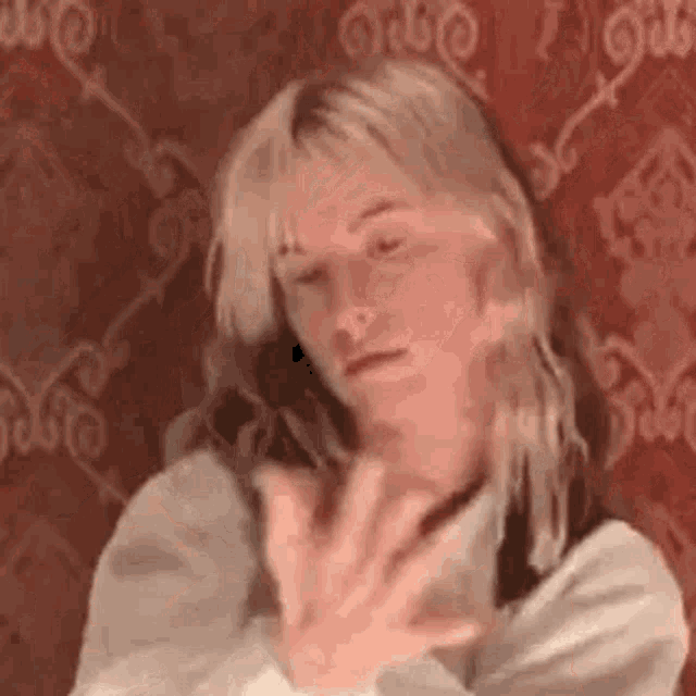 a woman with long blonde hair is sitting in front of a red wall and covering her face with her hands .
