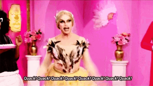 a drag queen in a gold dress is standing in front of a pink wall and flowers .